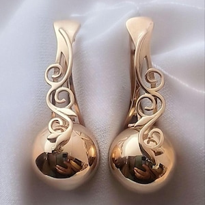 Puede incluir: Un par de pendientes dorados. Cada pendiente presenta una base esférica lisa y una parte superior curva y ornamentada con un diseño en espiral en forma de hoja. Los pendientes reflejan el entorno, creando un efecto de espejo.