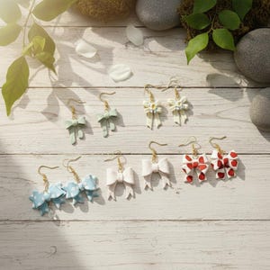 May include: A collection of handmade bow earrings in various colors, including light blue with white polka dots, white, red with white hearts, and mint green. Each earring features a gold-colored hook.