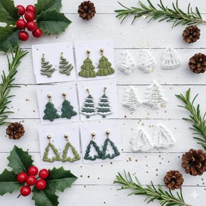 Könnte beinhalten: Eine Sammlung von Weihnachtsbaum-Ohrringen in verschiedenen Grüntönen, präsentiert auf weißen Karten. Die Ohrringe weisen unterschiedliche Designs auf, darunter massive, umrissene und geschichtete Baumformen. Das Bild ist mit Tannenzapfen, Stechpalmen und Rosmarinzweigen verziert.