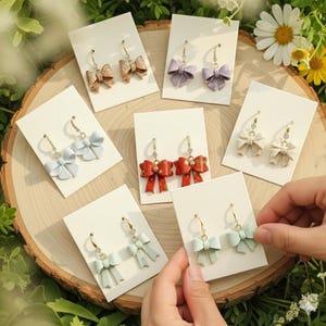 May include: A collection of bow-shaped earrings in various colors, including light blue, red, and beige, displayed on white cards. The earrings have gold-colored hoops and are arranged on a wooden surface, showcasing a variety of styles.