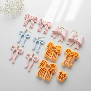May include: A selection of bow-shaped earrings and cookie cutters in various colours. The earrings are in pink and light blue, with gold-coloured accents. The cookie cutters are in orange. The bows have a heart-shaped centre.