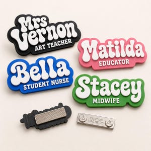 May include: A collection of colorful name badges with white text. The badges include names like "Mrs. Vernon" and "Bella", with job titles such as "Art Teacher" and "Student Nurse". A separate magnetic backing and a "Caution Magnetic" badge are also shown.