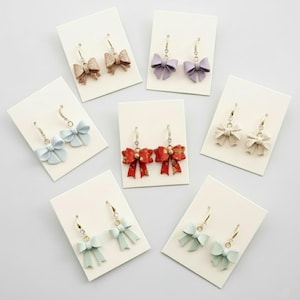 May include: A collection of bow-shaped earrings in various colors, including red, lavender, and light blue, displayed on white cards. The earrings have gold-colored hooks and some feature sparkling accents. The bows have a textured appearance.
