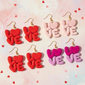 Könnte beinhalten: Sechs Paar Ohrringe in Rot-, Rosa- und Lilatönen. Jeder Ohrring zeigt das Wort "LOVE" in Herzform, mit goldfarbenen Ohrhaken. Die Ohrringe werden auf einem rosa Hintergrund mit verstreuten herzförmigen Konfetti präsentiert.