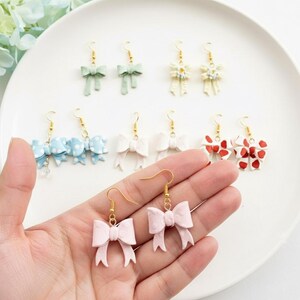 May include: A collection of handmade bow earrings in various colors, including light pink, mint green, and white. Some earrings feature polka dots or floral accents. The earrings are displayed on a white plate, showcasing their delicate designs and gold-colored hooks.