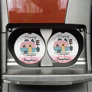 May include: Two round car coasters with a pink and teal camper design. The coasters have the words "Happy Camper Josephine" printed on them. The coasters are in a car's cup holders.