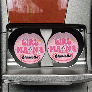 May include: Two pink car coasters with the text "GIRL MAMA" in a retro font, with the name "Charlotte" below. The coasters have a floral and lightning bolt design.
