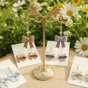 May include: A gold-colored earring display stand holding several pairs of bow-shaped earrings. The earrings are in various colors, including tan, lavender, and light blue. Each earring has a crystal accent. The background features greenery and white flowers.