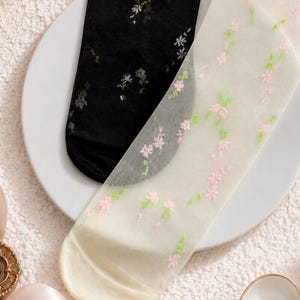 May include: Two pairs of sheer socks, one black and one off-white, each decorated with delicate floral embroidery. The black sock has white and grey flowers, while the off-white sock features pink and green blossoms. The socks are displayed on a white plate.
