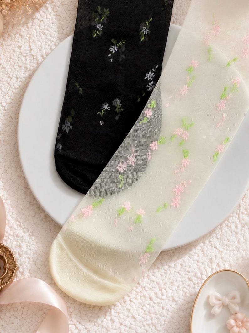 May include: Two pairs of sheer socks, one black and one off-white, each decorated with delicate floral embroidery. The black sock has white and grey flowers, while the off-white sock features pink and green blossoms. The socks are displayed on a white plate.