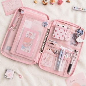 Könnte beinhalten: Ein rosa Ringbuch-Organizer mit Reißverschluss, Sechs-Ring-Mechanik aus Metall, mehreren transparenten Innentaschen und elastischen Stiftschlaufen, gefüllt mit Schreibwaren wie Stiften, einer Schere, Aufklebern und Karten mit der Aufschrift Flora.