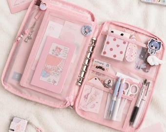 May include: A pink zipped binder organiser featuring a six-ring metal mechanism, multiple clear interior pockets, and elastic pen loops. The organiser is filled with stationery items including pens, scissors, stickers, and cards labelled Flora.