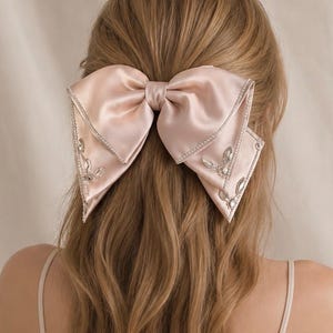 May include: A large, pale pink satin hair bow featuring a smooth, lustrous finish. The edges of the bow are embellished with delicate silver-toned trim and clear, faceted crystal accents arranged in a leaf-like pattern on the two hanging tails.