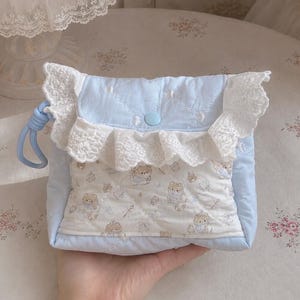 Puede incluir: Un bolso acolchado de color azul claro con un borde de encaje blanco en la solapa y un panel central decorado con un patrón repetitivo de pequeños ositos de peluche marrones. El bolso incluye un cierre de botón a presión y una correa de muñeca.