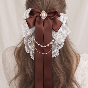 May include: A dark brown satin hair bow featuring a central oval faux pearl embellishment with a gold-toned frame. The bow is layered over white floral lace and accented with two delicate gold-toned chains draped with small white faux pearls.