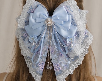 May include: A large hair bow featuring light blue satin ribbon layered over blue and purple floral embroidered lace with white scalloped lace trim. The centre is adorned with a pearl and crystal embellishment with dangling pearl and crystal chains.