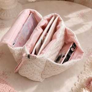 May include: A quilted white fabric bag with small grey polka dots and a soft pink interior lining. The bag features multiple open compartments containing a book with a light-coloured cover and a small pink digital camera with a black lens.