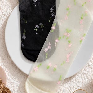 May include: A pair of sheer socks, one black and one off-white, both decorated with floral embroidery. The black sock has white and green flowers, the off-white sock has pink and green. These socks provide a delicate, elegant touch.