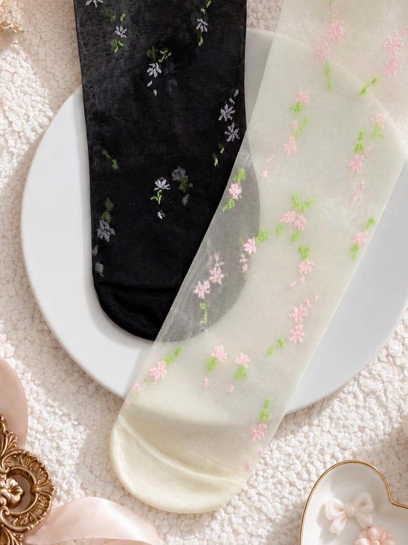 May include: A pair of sheer socks, one black and one off-white, both decorated with floral embroidery. The black sock has white and green flowers, the off-white sock has pink and green. These socks provide a delicate, elegant touch.