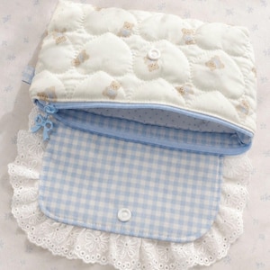 Puede incluir: Un estuche acolchado de tela blanca con un patrón repetitivo de pequeños ositos de peluche. El estuche tiene una cremallera azul claro con un tirador en forma de lazo y una solapa con estampado de cuadros vichy azul y blanco, con encaje blanco.