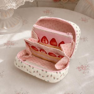 Peut inclure: Une trousse de toilette en tissu matelassé avec un extérieur crème orné d'un petit motif de fraises. L'intérieur est doublé d'un tissu rose à petites étoiles et comprend un compartiment central zippé décoré d'un motif de tranches de fraise.