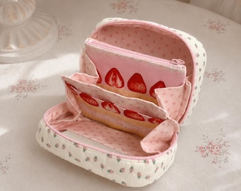 May include: A quilted fabric cosmetic bag with a cream exterior featuring a small strawberry print. The interior is lined with pink fabric patterned with tiny stars and features a central zippered compartment decorated with a strawberry slice print.