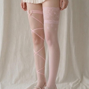 May include: A pair of light pink leg accessories consisting of one thigh-high stocking with a lace top and floral pattern, and one leg wrap featuring thin crisscross straps attached to a circular ring, paired with light pink pointed-toe flats.