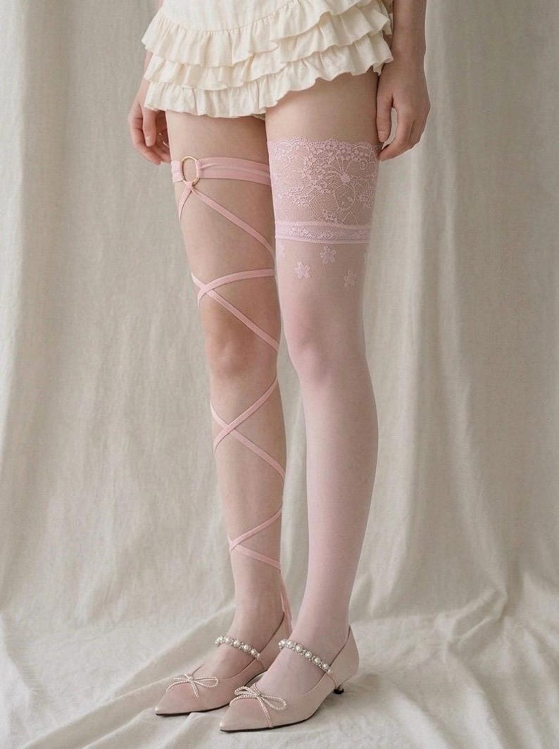 May include: A pair of light pink leg accessories consisting of one thigh-high stocking with a lace top and floral pattern, and one leg wrap featuring thin crisscross straps attached to a circular ring, paired with light pink pointed-toe flats.
