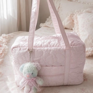 Op de afbeelding: Een lichtroze gewatteerde reistas met een all-over hartjespatroon, een ritssluiting aan de bovenkant en twee schouderbanden. Een kleine, pluizige, veelkleurige pastelkleurige teddybeer-sleutelhanger met een roze lint is aan een ring bevestigd.