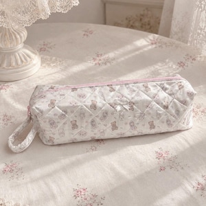 Puede incluir: Un estuche rectangular acolchado de tela blanca con un patrón repetitivo de pequeños osos de peluche marrones. El estuche tiene una cremallera rosa, un pequeño lazo de cinta blanca en la parte delantera y un asa de tela a juego.
