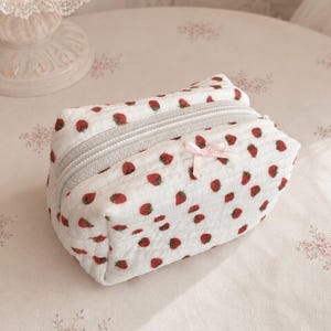 May include: A rectangular quilted pouch made of white fabric featuring a repeating pattern of small red strawberries. The pouch has a light grey zip closure along the top and a small, pale pink ribbon bow attached to the front centre.