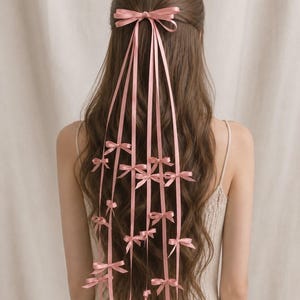 Op de afbeelding: Een haaraccessoire met een grote roze satijnen strik aan de bovenkant, met meerdere lange, dunne roze satijnen linten die naar beneden vallen, elk versierd met verschillende kleinere roze satijnen strikjes die langs de linten zijn bevestigd.