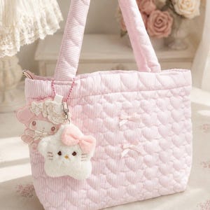 May include: A light pink quilted tote bag featuring a repeating heart pattern and thin white stripes. The bag has two top handles, two small decorative bows on the front, and two plush character charms attached to the side with a beaded chain.