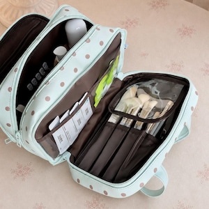 Peut inclure: Une trousse de toilette vert menthe clair à pois marron, dotée de multiples compartiments intérieurs, d'une doublure marron foncé et d'une section zippée en plastique transparent contenant plusieurs pinceaux de maquillage et divers petits objets.