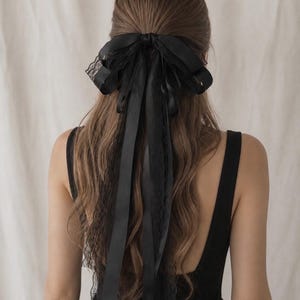 May include: A black hair accessory featuring a large bow made of satin ribbon and delicate lace trim. The bow is secured at the centre and has long, flowing ribbon tails that extend down the back, layered with matching black lace edges.