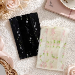 May include: Two rectangular pieces of sheer fabric, one black and one white, are presented on a textured white surface. The black fabric is embroidered with white floral sprigs, and the white fabric has pink and green floral embroidery. Roses and a teacup complete the scene.