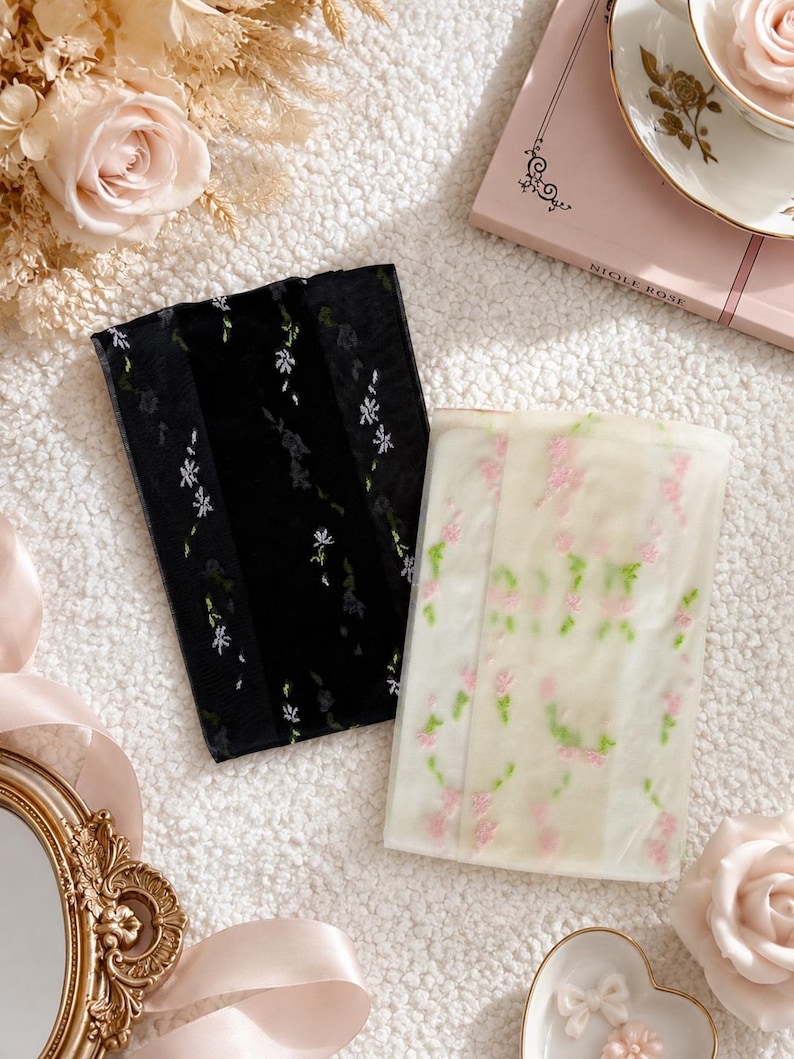 May include: Two rectangular pieces of sheer fabric, one black and one white, are presented on a textured white surface. The black fabric is embroidered with white floral sprigs, and the white fabric has pink and green floral embroidery. Roses and a teacup complete the scene.