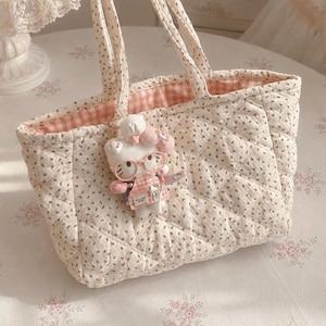 May include: A quilted tote bag featuring a cream fabric with a small dark floral print and two matching handles. The interior is lined with a pink and white gingham fabric. A plush white cat character charm wearing pink glasses is attached.