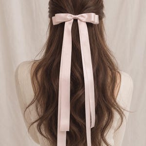 May include: A light pink satin ribbon hair bow with two long, trailing tails secured at the back of long, wavy brown hair. The smooth, lustrous fabric of the bow creates a soft, elegant appearance against the dark hair, with the ribbon tails hanging down.