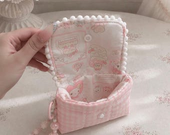 May include: A small, quilted, light pink and white gingham pouch with a flap closure featuring white pom-pom trim. The interior lining is white fabric with a repeating pattern of pink cakes and cartoon characters. A beaded wrist strap is attached.
