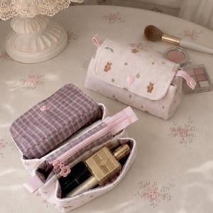 Könnte beinhalten: Zwei weiche, plüschige Kosmetiktaschen in hellem Rosa mit einem Muster aus kleinen braunen Bären und Blumen. Eine Tasche ist mit einem rosa Druckknopf verschlossen, während die andere geöffnet ist und ein lila-weiß kariertes Innenfutter zeigt.