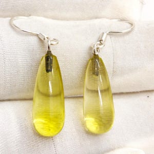 以下が含まれることがあります： 淡い黄色をした、しずく型のイヤリングのペア。イヤリングは銀色のフックと透明感のある外観をしています。イヤリングは白い表面に展示されています。