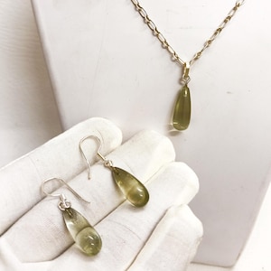 以下が含まれることがあります： オリーブグリーンの涙型の宝石をあしらった、ゴールドトーンのネックレスとイヤリングのセット。ネックレスは繊細なチェーンで、イヤリングはシンプルなフック留めです。ジュエリーは白い表面に展示されており、宝石の透明度を際立たせています。