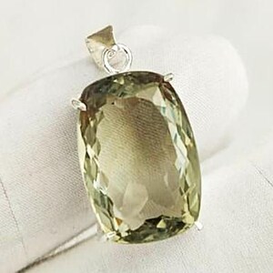 May include: A cushion-cut, pale olive-green gemstone pendant featuring a faceted surface and a silver-toned metal bail at the top. The stone displays a translucent quality with visible internal clarity and a rectangular shape with rounded corners.
