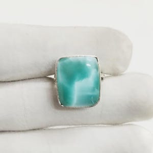 May include: A square-cut, light blue gemstone ring with a silver band. The gemstone has a marbled appearance with white and turquoise hues. The ring is set on a white surface.