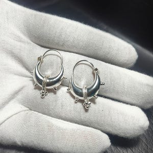 Könnte beinhalten: Ein Paar silberfarbene Creolen. Jeder Ohrring hat ein halbmondförmiges Design mit dekorativen Elementen, darunter kleine Kugeln und ein strukturiertes Band. Die Ohrringe werden auf einer weißen Oberfläche präsentiert.