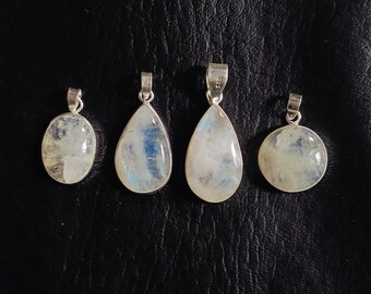 May include: Four silver pendants with iridescent moonstone cabochons. The pendants are in oval, teardrop, and round shapes. The moonstones have a milky white base with blue and gray internal patterns. The pendants are displayed on a dark background.