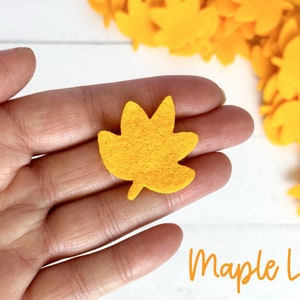 May include: A single yellow felt maple leaf held in a hand. The leaf is about 2 inches wide and 2 inches tall. The leaf has a slightly textured surface.