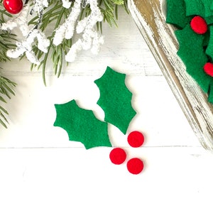May include: Green felt holly leaves and red felt berries on a white wooden surface.