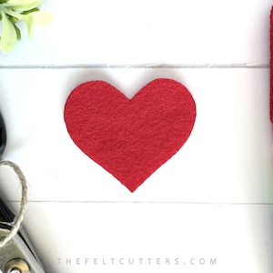 May include: A red felt heart shape, approximately 2 inches in diameter, on a white wooden background. The felt heart is a craft supply for making cards, decorations, and other projects.